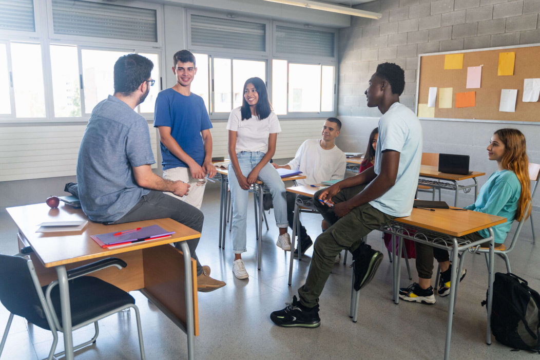 A group of students sitting and standing in a classroom, engaged in a casual discussion, with desks and a bulletin board in the background.