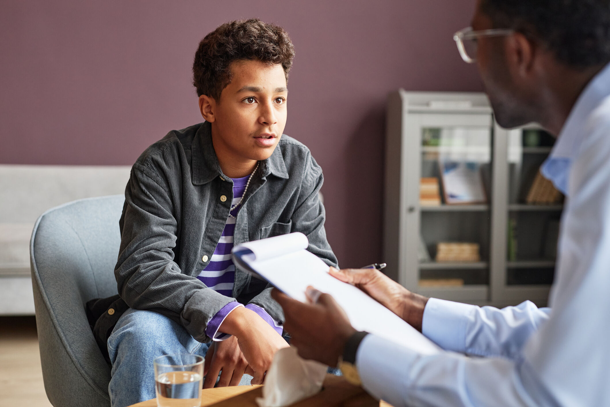 A young man sitting across from a professional who is holding a notepad, engaged in a focused conversation, possibly in a counseling or interview setting.