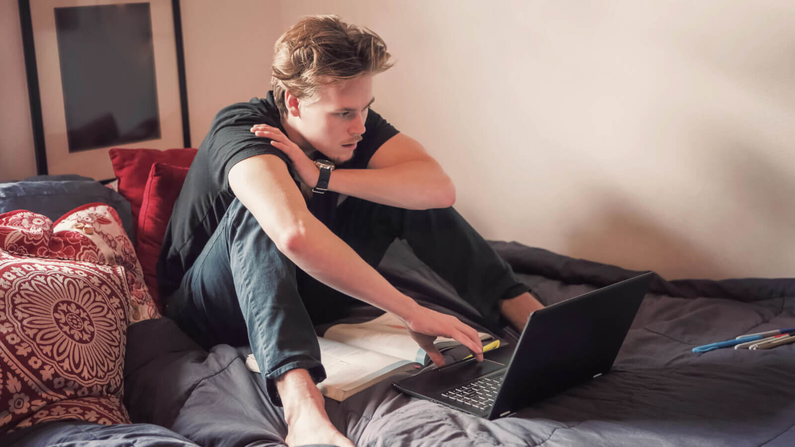 a student sits on a bed and does class work on a laptop