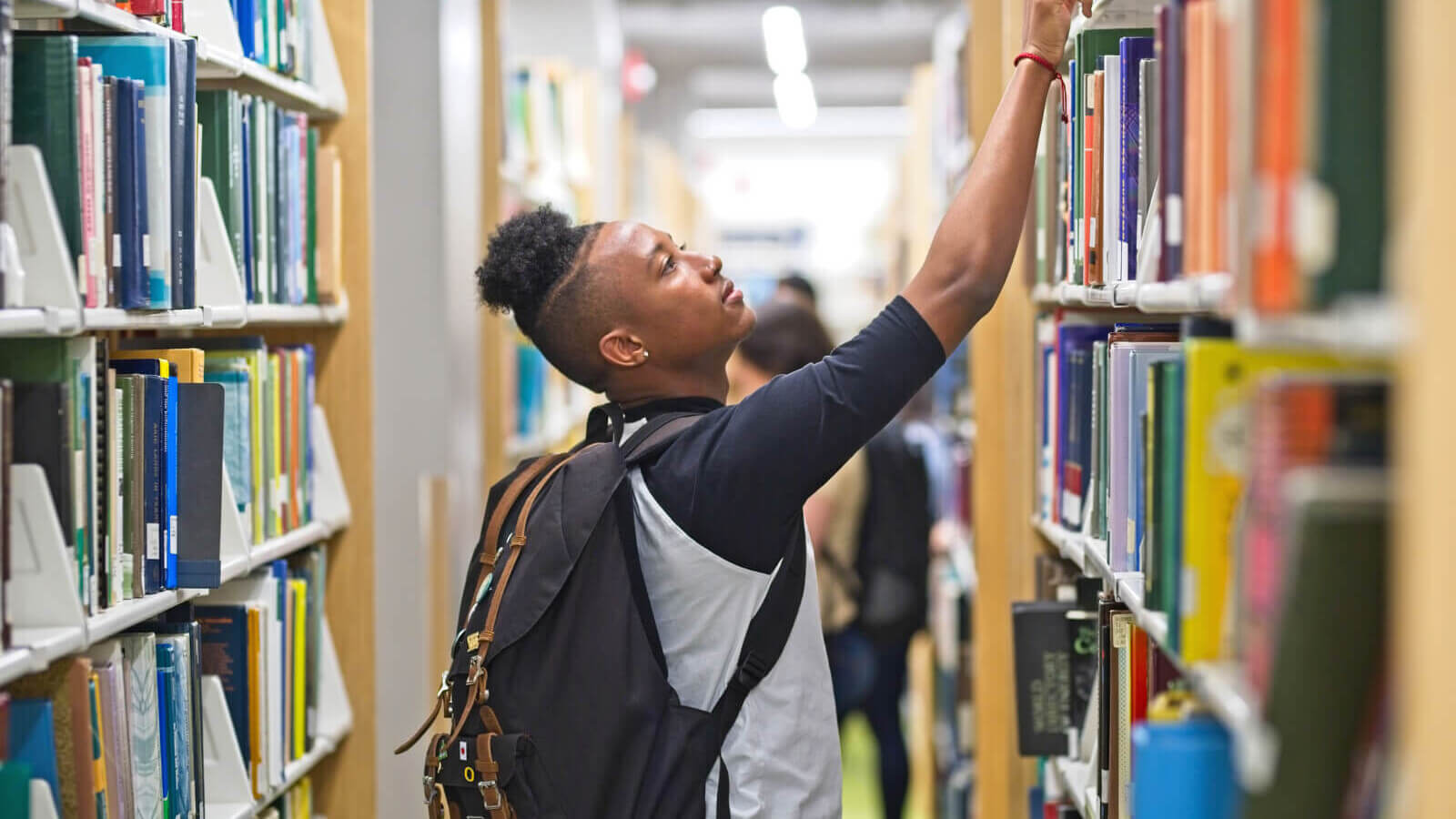 A male student in the library reaching the top shelf.