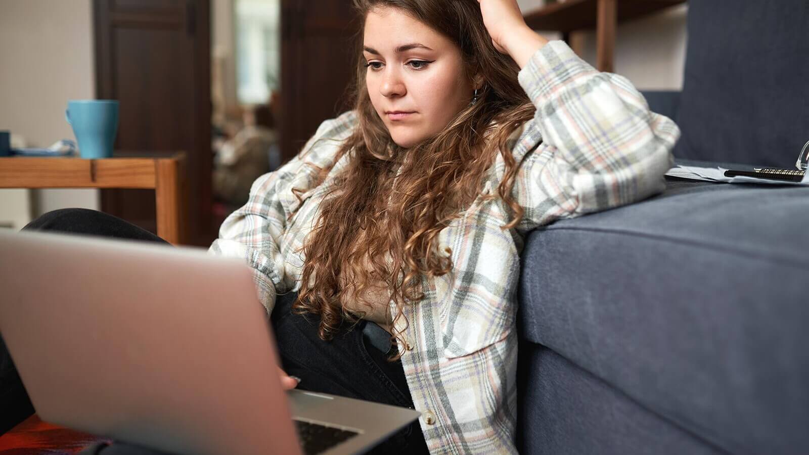 a student sits on the couch working on her laptop, looking stressed.